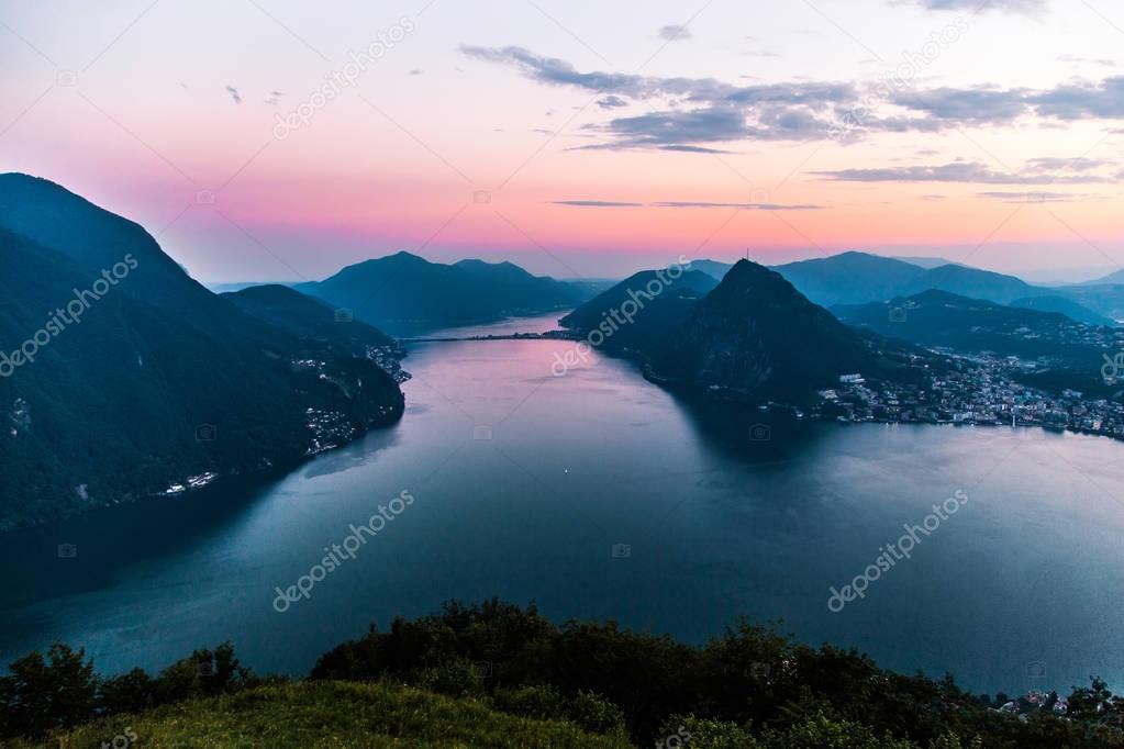 Aerial view of the lake Lugano surrounded by mountains and evening city Lugano on during dramatic sunset, Switzerland, Alps. Travel