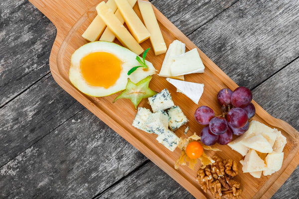 Cheese platter garnished with pear, honey, walnuts, grapes, carambola, physalis on cutting board on wooden background. Snacks and Wine appetizers set. Top view