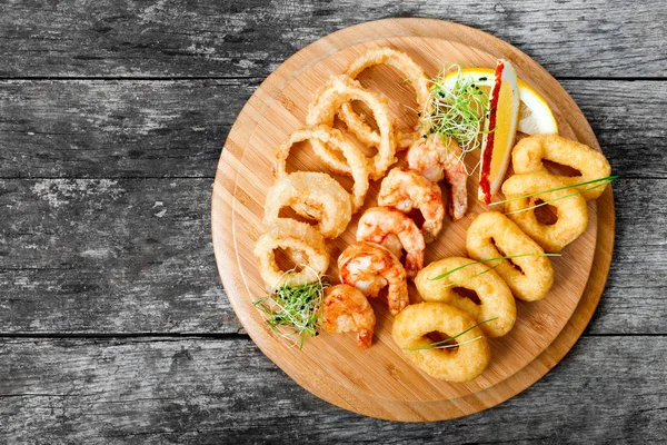 Piatto di pesce con anelli di calamari fritti, gamberi e anelli di cipolla decorati con limone sul tagliere su sfondo di legno. Antipasti mediterranei. Vista dall'alto — Foto Stock