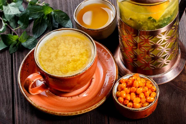 Tee mit Sanddorn in Tasse mit Untertasse, serviert mit Vintage-Teekanne, frischen Beeren, Honig, Minze auf dunklem Holzgrund. gesunde Getränke — Stockfoto