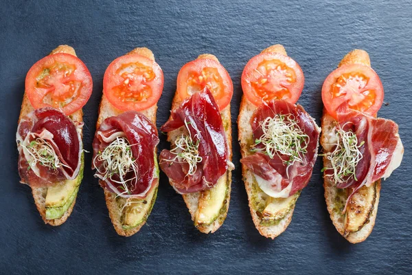 Aperitivo bruschetta com prosciutto, tomate, abobrinha em pão ciabatta em pedra ardósia fundo fechar. Deliciosos lanches, sanduíches, crostini, antipasti na festa ou horário de piquenique. Vista superior — Fotografia de Stock