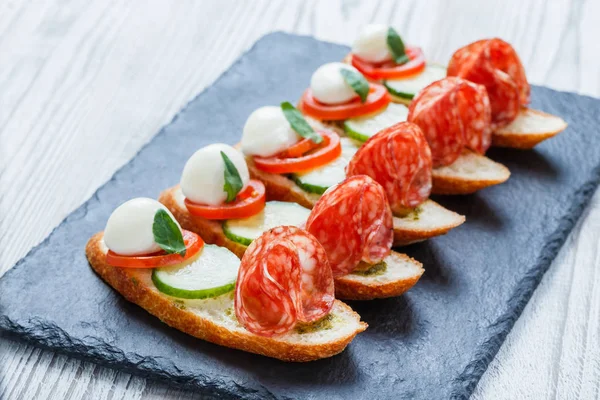 Sanduíches Caprese com tomate, queijo mussarela, manjericão, salame no pão ciabatta no fundo de ardósia de pedra perto. Deliciosos petiscos aperitivos, bruschetta, crostini, antipasti. Vista superior — Fotografia de Stock