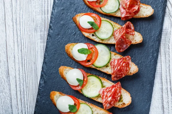 Sanduíches Caprese com tomate, queijo mussarela, manjericão, salame no pão ciabatta no fundo de ardósia de pedra perto. Deliciosos petiscos aperitivos, bruschetta, crostini, antipasti. Vista superior — Fotografia de Stock