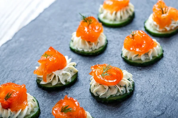 Appetizer canape with salmon, cucumber and cream cheese on stone slate background close up. Delicious snacks, sandwiches, crostini, brushetta, antipasti on party or picnic time. Top view