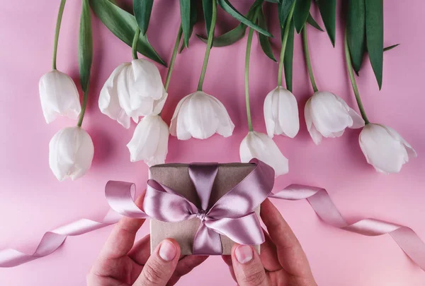 Manos masculinas sosteniendo un regalo con cintas sobre fondo rosa con tulipanes blancos. Piso tendido, vista superior — Foto de Stock