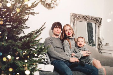 Noel ailesi ve Noel ağacının yanında gülümseyen bir bebek. Noel ağacı ve hediye kutularıyla dekore edilmiş oturma odası. Işık rahat bir ortam sağlıyor. Yeni yıl tatili
