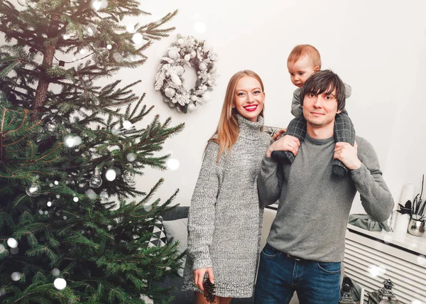 Різдвяна сім'я з дитиною посміхається біля ялинки. Вітальня, прикрашена ялинкою та подарунковими коробками, надає світлу затишну атмосферу. Новорічні свята — стокове фото