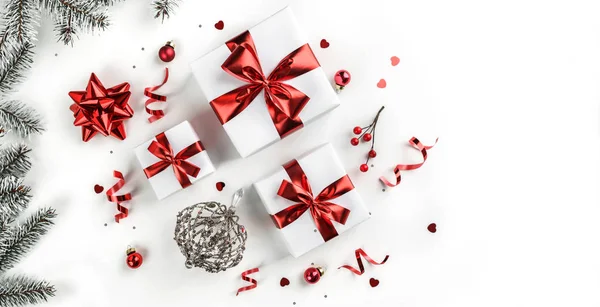 Noel ağacı dalları, kırmızı kurdeleli hediye kutuları, kırmızı süslemeler, parıltılar ve beyaz arka planda konfeti. Noel ve yeni yıl tebrik kartı, kış tatili. Düz yatış, üst görünüm