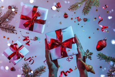Bayanların elinde renkli parlak neon rengi parlak mavi ve mor arka planda uçan Noel süslemeleri, köknar dalları ve konfetileri olan hediye kutusu var. Noel ve Yeni Yıl tatili, Bokeh, ışık. Seçici odak