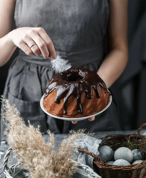 Női kéz kezében csokoládé Húsvéti torta fehér asztalon díszített tavaszi virágok és fonott kosár tojással. Kellemes húsvéti ünnepeket, szelektív fókuszt — Stock Fotó