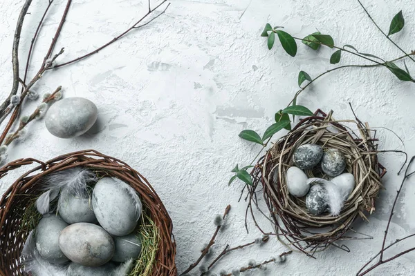 Modern Easter eggs in wicker basket on white textured background with nest, feathers and spring flowers. Happy Easter holiday, top view, flat lay