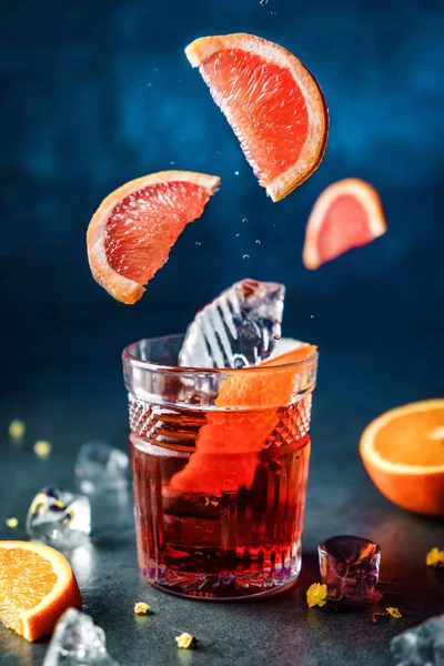 Cocktail de toranja fresco com laranja e gelo em vidro sobre fundo azul escuro. Estúdio tiro de bebida em movimento de congelamento, voando fatias de toranja, baixas em respingo líquido. Bebida fria de verão e coquetel — Fotografia de Stock