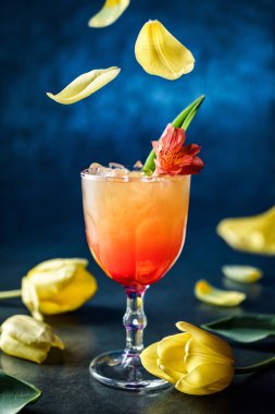 Koyu mavi arka planda şarap kadehinde çiçekler olan taze tropikal kokteyl. Stüdyo içkisi dondurucu hareketle, sarı lale yaprakları havada uçuşuyor. Soğuk yaz içeceği ve kokteyl, seçici odaklanma