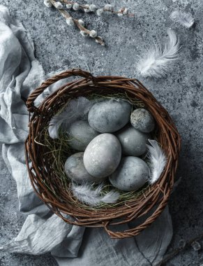 Hasır sepette paskalya yumurtaları, mavi arka planda tüyler ve bahar çiçekleri. Mutlu Paskalyalar, üst manzara, düz yatak