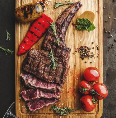 Orta pişmiş ızgara tomahawk eti mi yoksa ahşap kesme tahtasında ızgara sebze ve koyu arkaplanda şarap kadehi mi? Üst görünüm