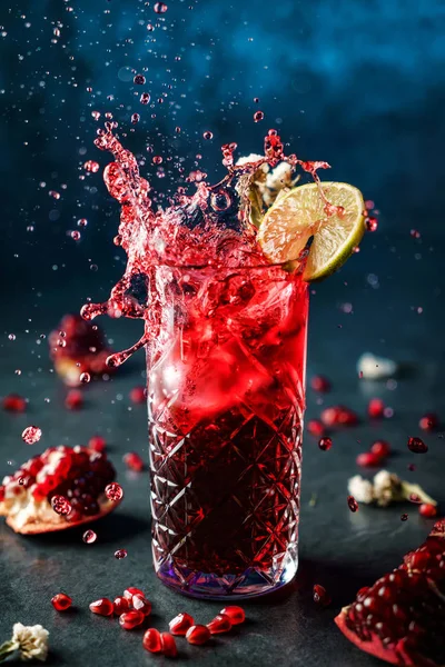 Fresh pomegranate cocktail with lime in drink glass on dark blue background. Studio shot of drink in freeze motion, splash and drops in flying. Summer cold drink and cocktail — Stock Photo, Image