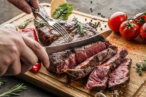 Ruce krájené grilované tomahawk maso střední vzácné nebo rib oko steak na dřevěné řezací desce s grilovanou zeleninou na tmavém pozadí, zblízka — Stock fotografie