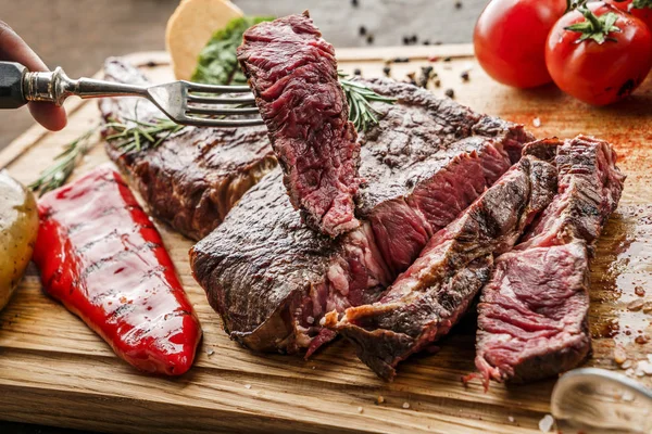 Ruce krájené grilované tomahawk maso střední vzácné nebo rib oko steak na dřevěné řezací desce s grilovanou zeleninou na tmavém pozadí, zblízka — Stock fotografie