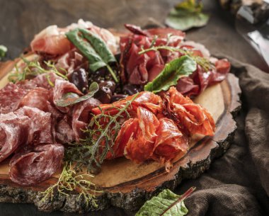 Antipasto tabağı prosciutto, jambon, salam, fesleğen ve koyu ahşap zemin üzerine zeytin ile süslenmiş soğuk et tabağı. Et aperatifi, şarap seti, yakın çekim.