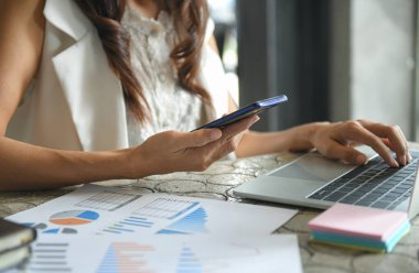 Bir iş kadını bilgi almak için cep telefonu kullanır. Akıllı telefonu tutar ve elini dizüstü bilgisayarın klavyesine koyar..
