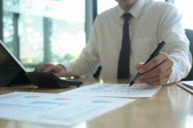 Genç işadamı bir grafikte verileri kontrol ediyor ve bir ofis masasında dizüstü bilgisayar kullanıyor. Grafikteki kalem noktasını kullanıyor ve diğer elini de klavyeye koyuyor..