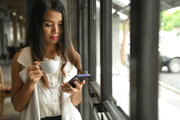 コーヒーカップを手にした白いドレス姿のアジア人女性が携帯電話を使っていて カフェの自然光が差し込む窓側に立っていた — ストック写真