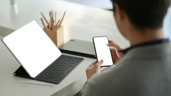 Young businessman hold a blank screen smartphone in hand and a blank screen laptop on the desk.