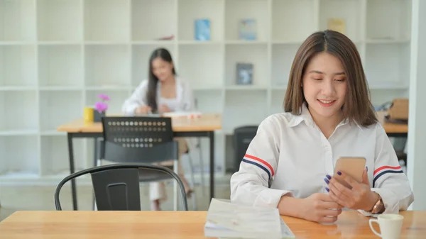 白いシャツを着た若い女性の肖像画は 現代のオフィスでスマートフォンを使用しています 後ろにはもう一人の女の子が座っていた — ストック写真