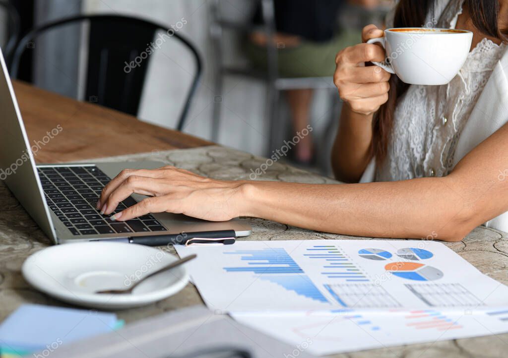 Female businessmen are eating coffee and using laptop. She works between the coffee brake, graphs on the desk.