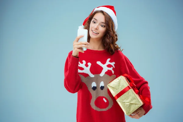 Dívka oblečená do Santa klobouku s vánočním dárkem a telefonem — Stock fotografie