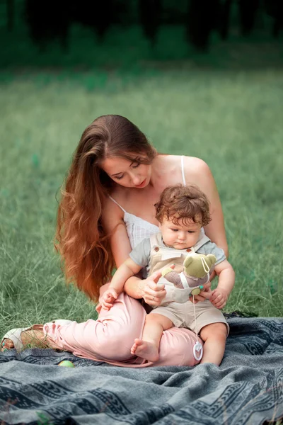 Jovem mãe bonita sentada com seu filho pequeno contra a grama verde. Mulher feliz com seu menino em um dia ensolarado de verão. Família caminhando no prado . — Fotografia de Stock
