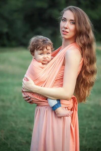 Jovem mãe bonita abraçando seu pequeno filho criança contra a grama verde. Mulher feliz com seu menino em um dia ensolarado de verão. Família caminhando no prado . — Fotografia de Stock