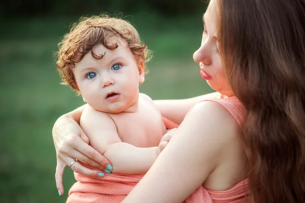 Jovem mãe bonita abraçando seu pequeno filho criança contra a grama verde. Mulher feliz com seu menino em um dia ensolarado de verão. Família caminhando no prado . — Fotografia de Stock