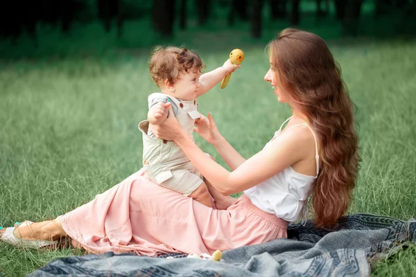 Die junge schöne Mutter sitzt mit ihrem kleinen Sohn im grünen Gras. glückliche Frau mit ihrem kleinen Jungen an einem sonnigen Sommertag. Familie spaziert auf der Wiese. — Stockfoto