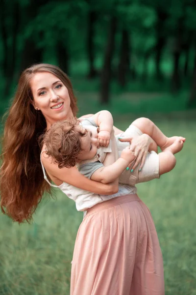 Jovem mãe bonita abraçando seu pequeno filho criança contra a grama verde. Mulher feliz com seu menino em um dia ensolarado de verão. Família caminhando no prado . — Fotografia de Stock