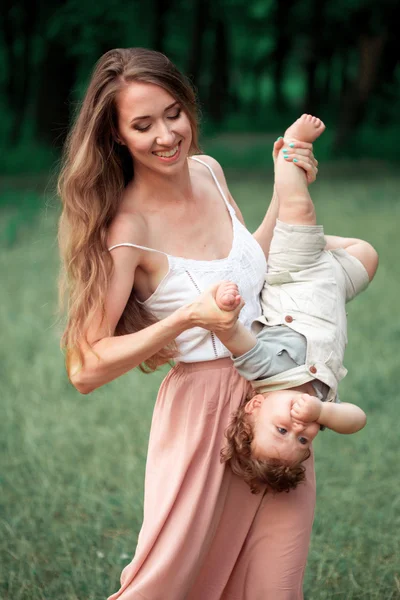 Jovem mãe bonita abraçando seu pequeno filho criança contra a grama verde. Mulher feliz com seu menino em um dia ensolarado de verão. Família caminhando no prado . — Fotografia de Stock