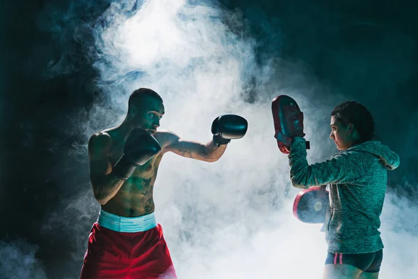 Guapo boxeador afroamericano con torso desnudo está practicando golpes con un compañero en el club de lucha —  Fotos de Stock