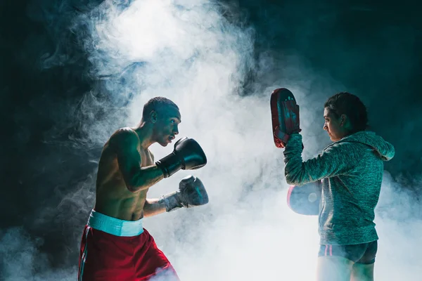Guapo boxeador afroamericano con torso desnudo está practicando golpes con un compañero en el club de lucha —  Fotos de Stock