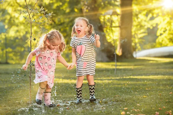 Las niñas rubias lindas en botas de goma jugando con salpicaduras de agua en el campo en verano — Foto de Stock