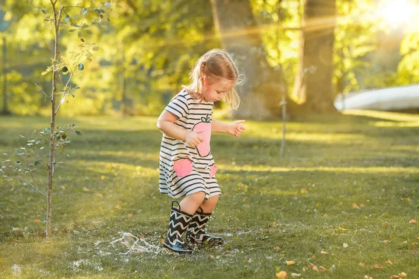 Ładny blond dziewczynka w gumowe buty, gra z wodą rozpryskową na polu w lecie — Zdjęcie stockowe