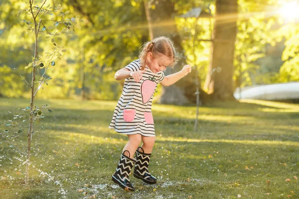 La linda niña rubia en botas de goma jugando con salpicaduras de agua en el campo en verano — Foto de Stock