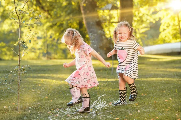 As meninas loiras bonitos em botas de borracha brincando com salpicos de água no campo no verão — Fotografia de Stock