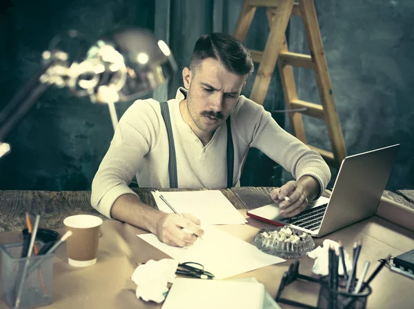 Красивий елегантний чоловік сидить за домашнім столом, працює і використовує ноутбук під час куріння сигарет — стокове фото
