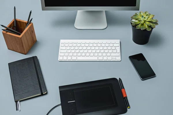 A mesa cinza com laptop, bloco de notas com folha em branco, vaso de flor, estilete e tablet para retoque — Fotografia de Stock