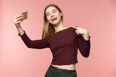 Şirin esmer kız bir selfie alır. O yaygın gülümsüyor. Uzun dalgalı esmer saçları. Güzellik kız pembe arka plan üzerinde.