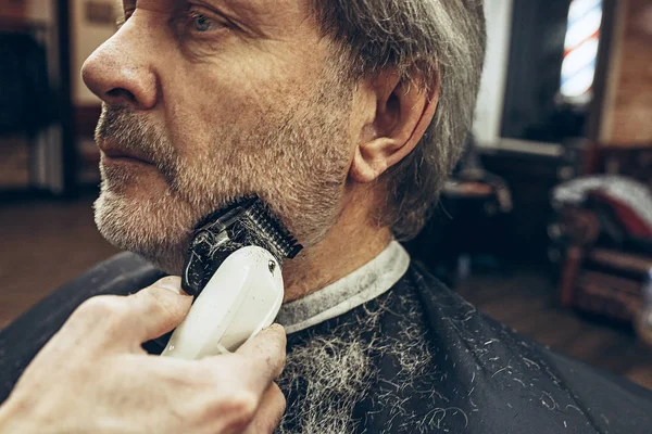 Close-up vista lateral retrato de belo homem caucasiano barbudo sênior recebendo barba grooming na barbearia moderna . — Fotografia de Stock