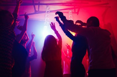 Silüetli bir kalabalık parti arka planındaki renkli neon ışığa karşı ellerini kaldırıyor.