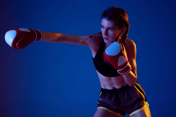 Fit caucasian woman in sportswear boxing on blue studio background in neon light — Stock Photo, Image