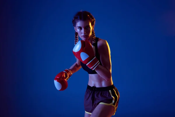 Fit caucasian woman in sportswear boxing on blue studio background in neon light — Stock Photo, Image