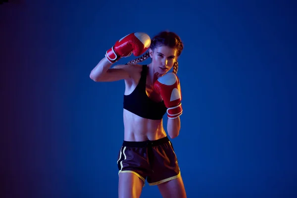 Fit caucasian woman in sportswear boxing on blue studio background in neon light — Stock Photo, Image